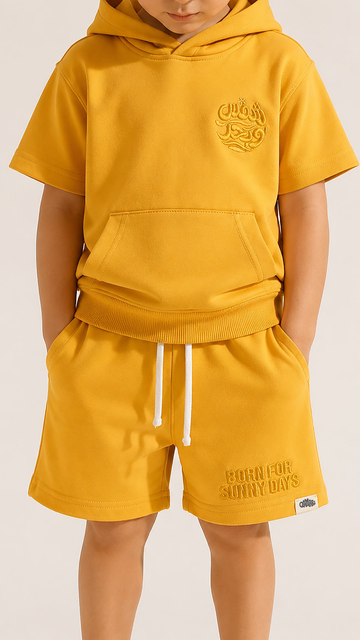 A child wearing a matching mustard yellow hoodie and shorts set with white drawstrings, photographed against a plain light beige studio background.