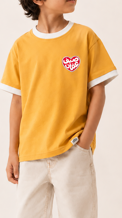 A child wearing a mustard yellow ringer T-shirt with white trim on the collar and sleeve cuffs, featuring a red and white heart graphic on the left chest. The child is also wearing light beige pants, set against a plain, warm off-white studio background.