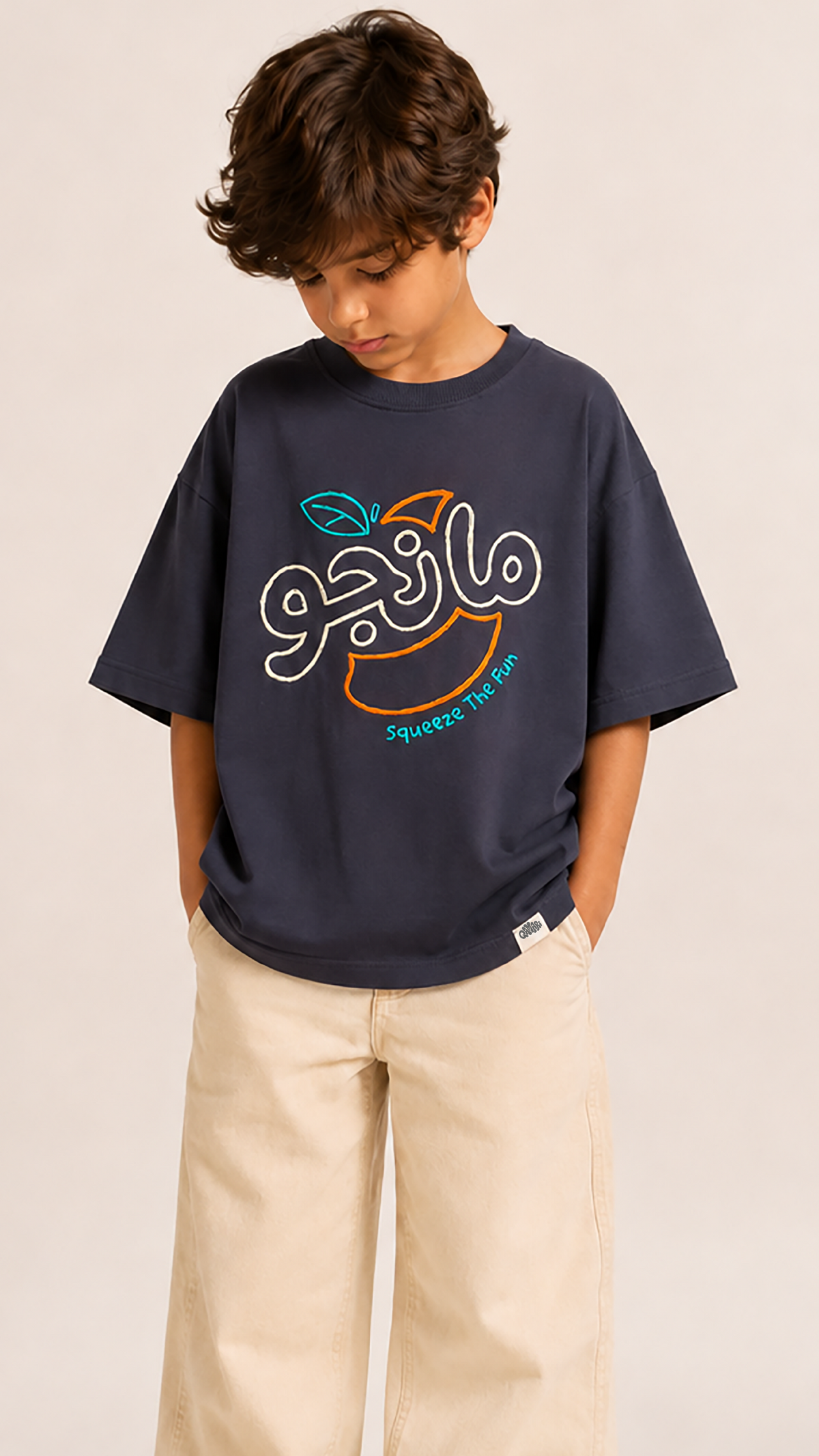 A young boy wearing a navy blue t-shirt with a colorful graphic print of an orange and Arabic script, paired with beige pants, standing against a light beige background.