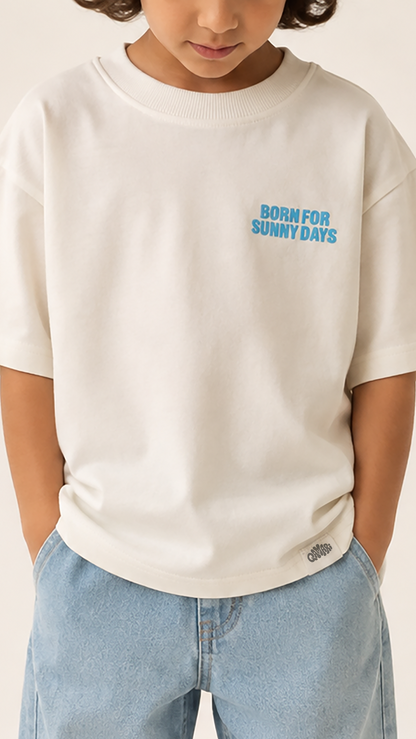 A child wearing an off-white short-sleeve t-shirt with 'BORN FOR SUNNY DAYS' printed in light blue on the left chest, paired with light blue jeans against a warm beige background.