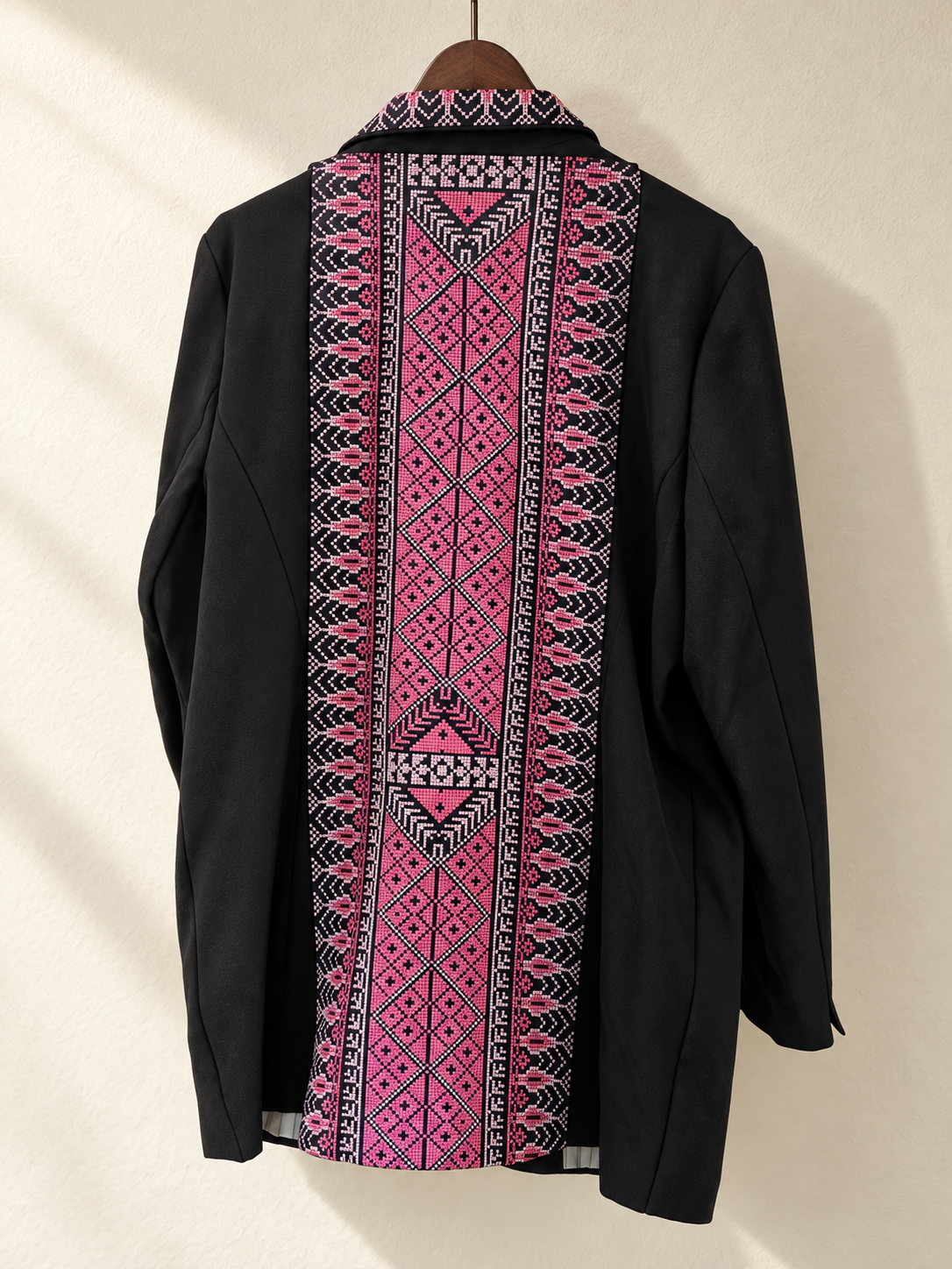 Back view of black blazer with a long pink geometric embroidered panel on a wooden hanger